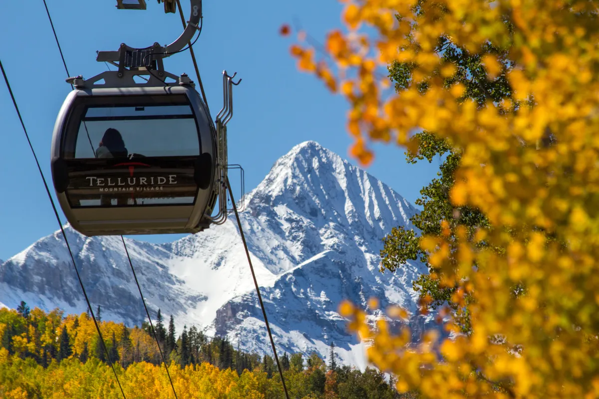 A gondola glides above a snow-capped mountain, with golden autumn leaves in the foreground, capturing both the adventure of skiing and scenic rides. The crisp landscape evokes the thrill of immediate mountain access.