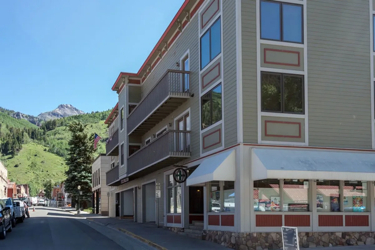 An energetic street-level view of a building facade with balconies looking out to the mountains, bustling with activity as people explore, shop, or dine. The mountain backdrop frames the scene, capturing both the urban vibrancy and natural beauty of Telluride.