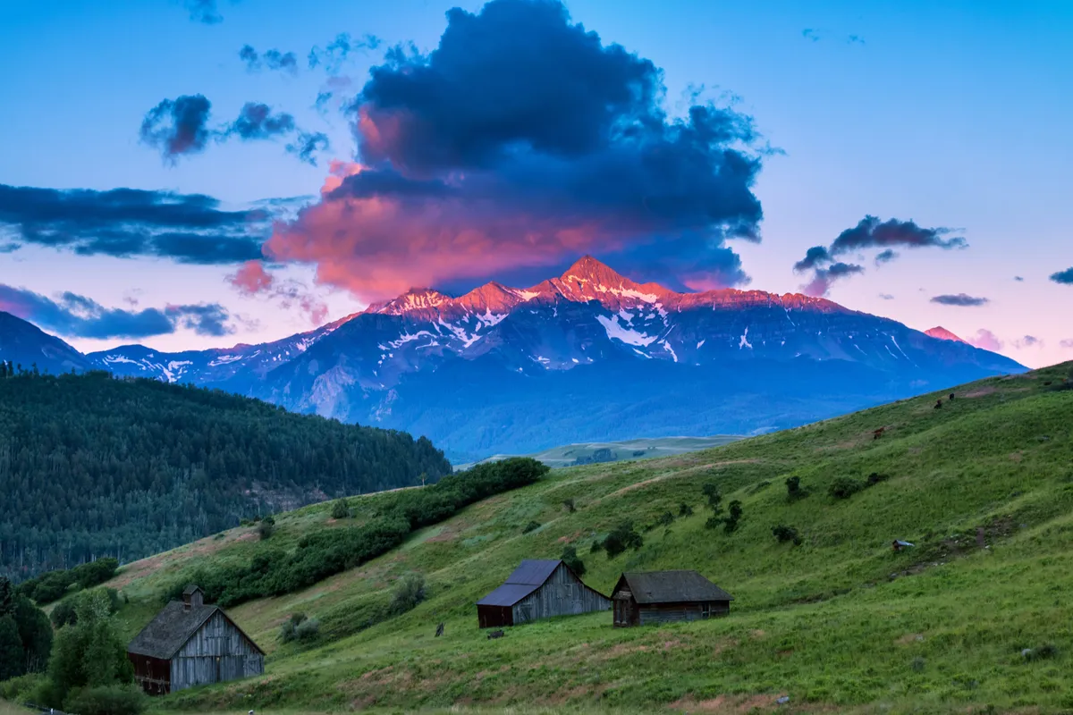 A peaceful exterior view highlighting rustic buildings nestled among greenery, with mountains and clouds in the background. It shows the surrounding nature and inviting entrance to the local area.