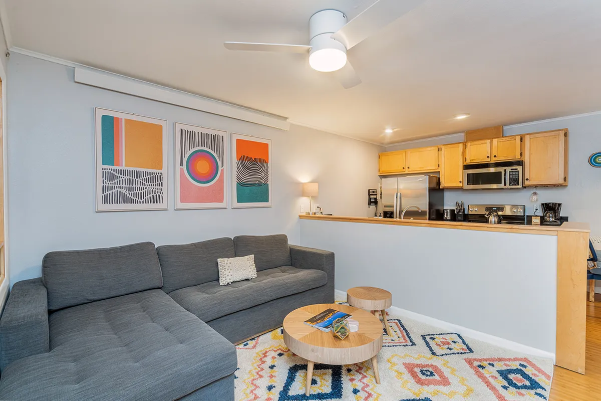 A modern, cozy living room with a gray sectional sofa, colorful wall art, a wooden coffee table, and a view of the adjacent kitchen. The scene is set for relaxed gatherings or quiet moments after a day outdoors.