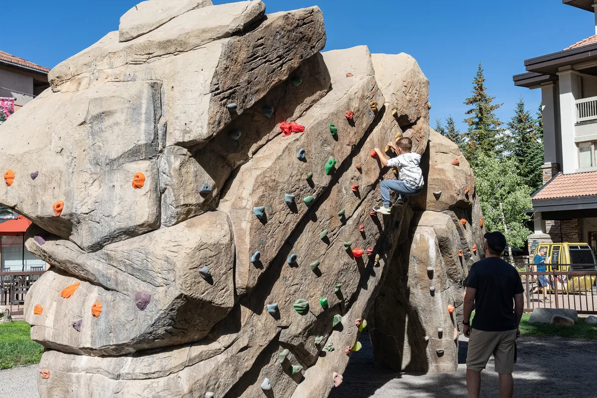 A child is climbing a rock structure at an outdoor playground while an adult observes closely, set against a backdrop of greenery and open space. The mood is lively and safe, emphasizing active outdoor play and parent-child bonding.