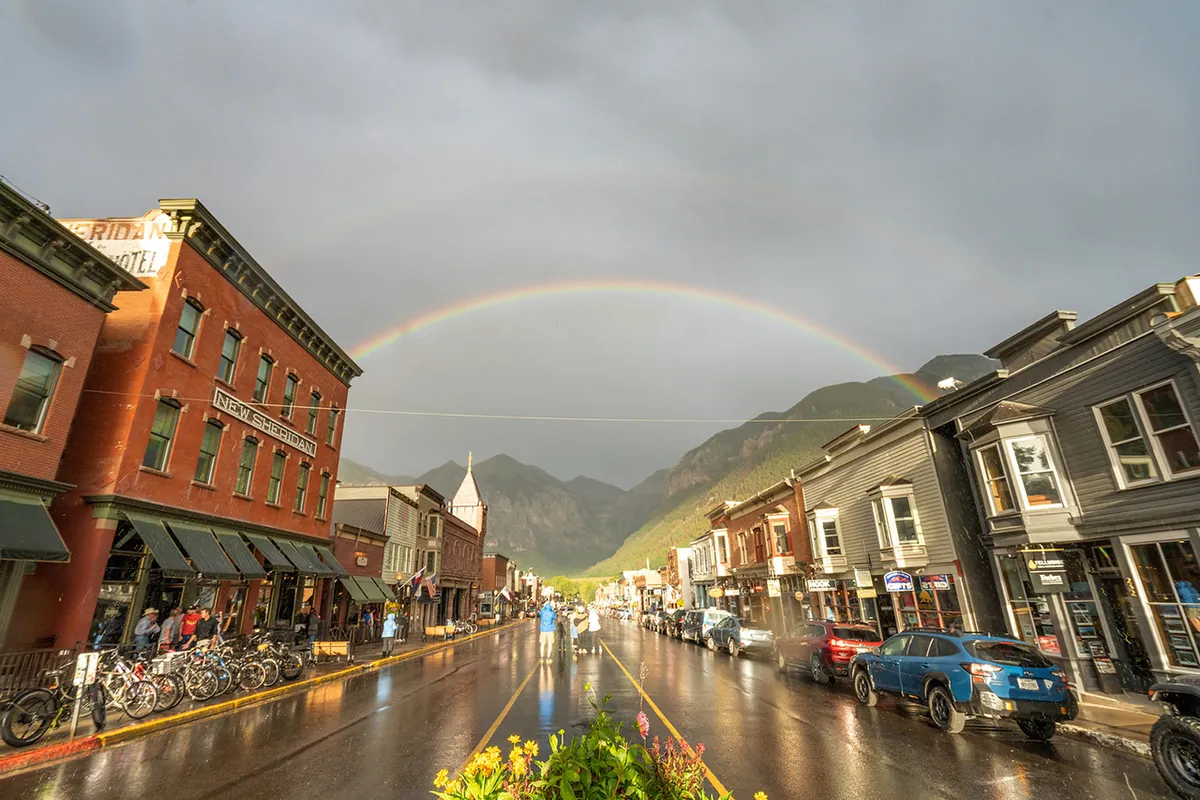 Historic downtown buildings line a wet street, with a vibrant rainbow arching overhead. People are walking and biking, and colorful details lend a cheerful, welcoming atmosphere to the scene.