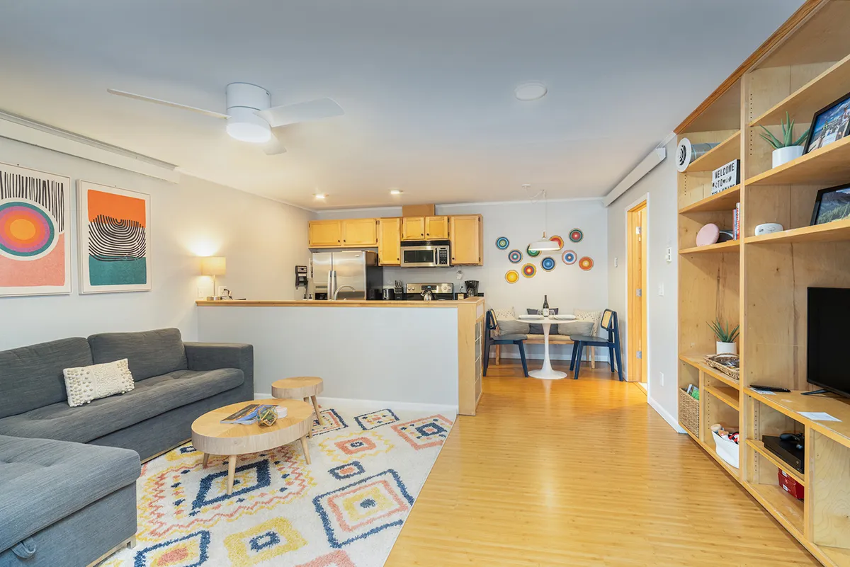 An inviting open-plan living space featuring a plush sofa, kitchen island, dining table, and vibrant artwork on the wall. The layout encourages relaxation, conversation, and easy movement between the living, dining, and kitchen areas.