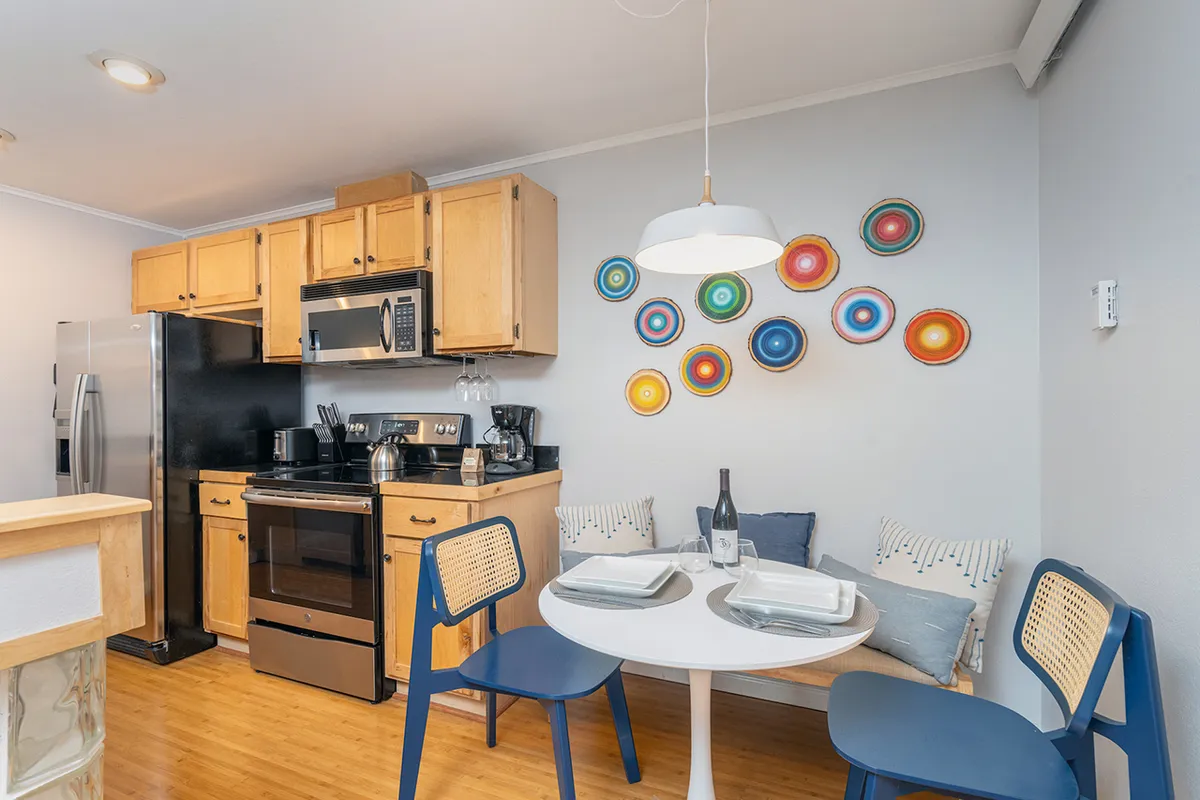The image showcases a bright, modern kitchen with stylish cabinets, a welcoming dining table set for a meal, and vibrant, colorful wall decor that adds warmth and character. The layout invites social gatherings and cozy meals, with plenty of natural light enhancing the cheerful, inviting atmosphere.