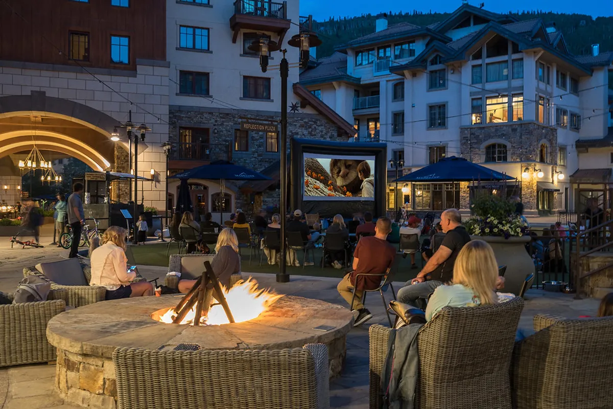 A warm, festive outdoor area featuring a fire pit, comfortable seating, and an outdoor movie screen, surrounded by nearby buildings. People gather and socialize in the inviting space, evoking a sense of community and evening relaxation.