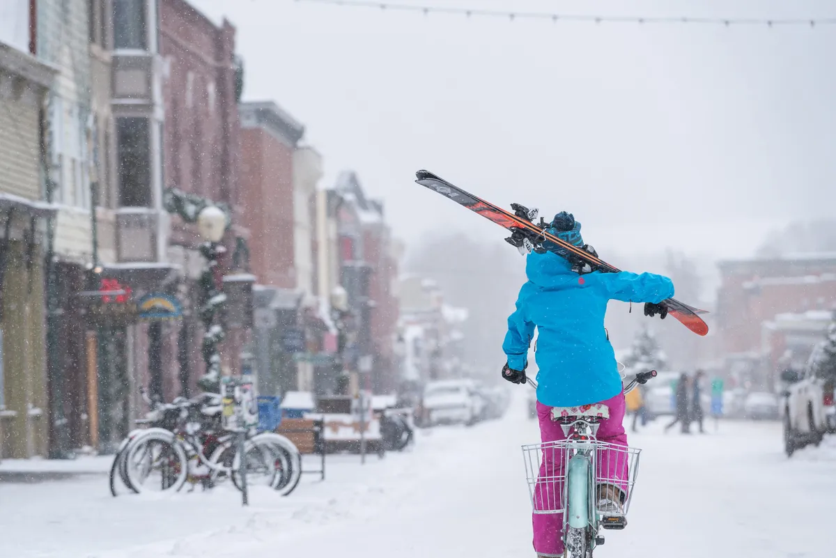 A lively winter street scene featuring a person cycling with skis overhead, surrounded by colorful storefronts and snow-covered roads. The setting is energetic, playful, and distinctly local, highlighting the car-free, adventure-centric lifestyle described in the section.