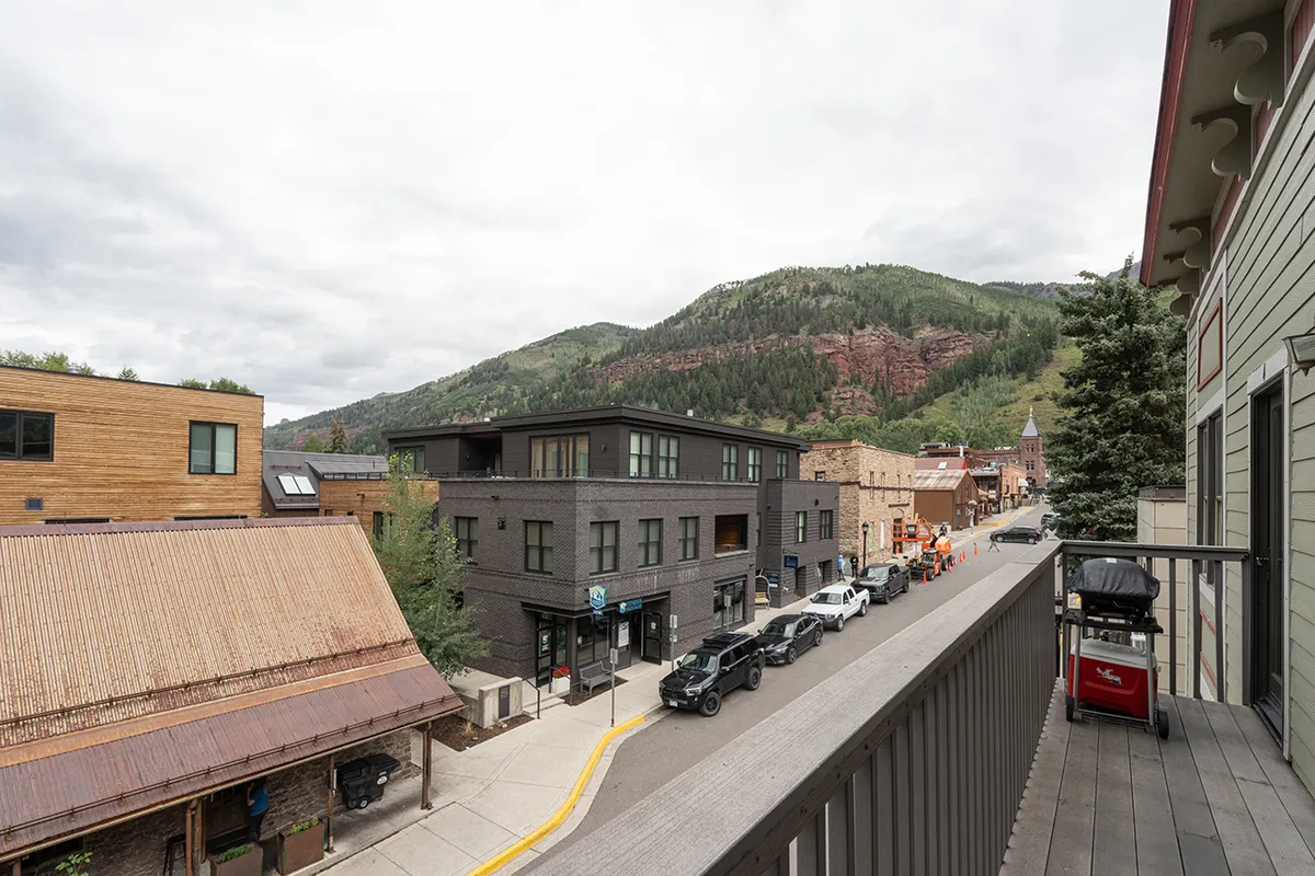 A tranquil street scene in Telluride featuring modern and historic buildings against a dramatic mountain backdrop. People can be seen strolling along the sidewalk, and several parked cars hint at the convenience and accessibility of the Main Street area. The image conveys the town's inviting atmosphere and proximity to local attractions.