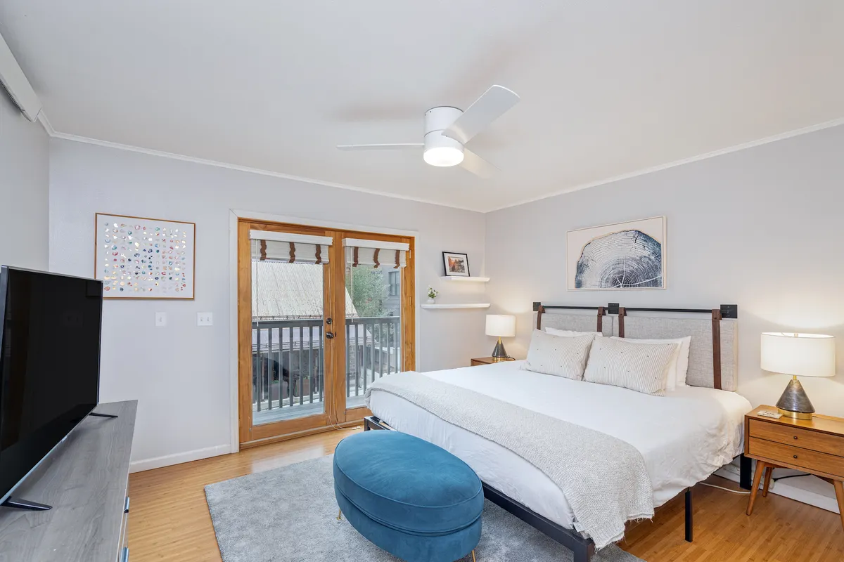 A serene master bedroom featuring a large, plush bed adorned with crisp linens. A blue ottoman sits at the foot of the bed, while double doors open out onto a private balcony, allowing soft natural light to fill the room. The space exudes comfort and relaxation, perfect for unwinding at the end of the day.