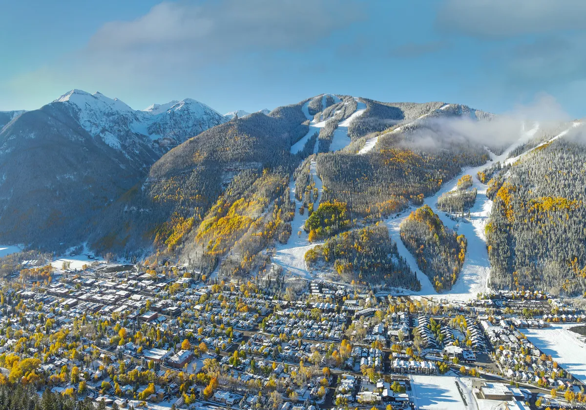 A panoramic view of snow-capped mountains, ski slopes, and a charming town dotted with colorful autumn trees. The landscape captures both winter sports excitement and the vibrant town atmosphere, ideal for exploration after outdoor activities.
